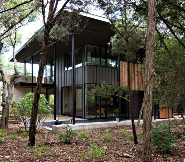 Exterior view of a contemporary multi-level house set among mature trees. The building features dark vertical cladding, extensive floor-to-ceiling glazing, slender dark structural posts, projecting roof planes, and a timber slatted privacy screen. A raised deck and glazed ground-level living spaces are visible through the trees.