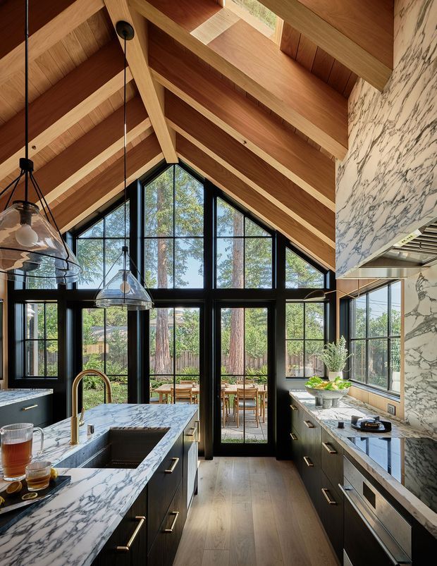 A dramatic, light-filled kitchen with a vaulted timber-lined ceiling, exposed timber beams and clerestory-like roof glazing. Black-framed, floor-to-ceiling windows and glazed doors open the room visually to a dining terrace and wooded garden. The kitchen features dark fixed cabinetry, extensive white marble surfaces with bold grey veining, brass-toned hardware and tapware, pendant lights, and a large island. A marble-clad rangehood feature forms a strong vertical element on the right.