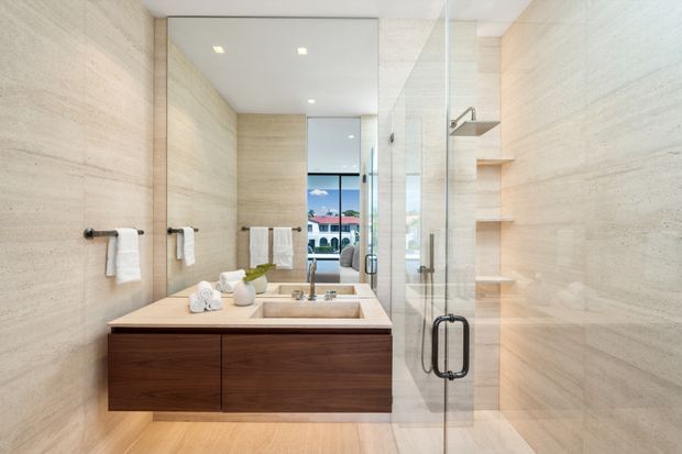 A bright contemporary bathroom with full-height pale stone wall and floor surfaces, a floating dark timber vanity with a pale stone integrated basin, wall-mounted chrome tapware, towel rails, and a frameless glass shower enclosure. Open recessed shelving is built into the shower wall, and a large mirror reflects the vanity and a glimpse of an adjacent room and exterior beyond.