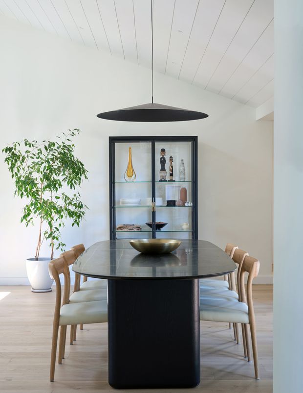 Bright, refined dining room with a large dark stone dining table and sculptural black pedestal base, surrounded by light upholstered timber dining chairs. A black glass-front display cabinet, decorative objects, a tall indoor plant, and a broad black pendant light sit against white walls and a white-lined pitched ceiling. Pale timber flooring creates a warm, clean contrast.