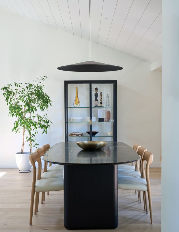 Bright contemporary dining room with a long dark stone dining table supported by a sculptural black pedestal base, surrounded by light timber dining chairs with pale upholstered seats. A black oval pendant light hangs from a white painted timber-lined vaulted ceiling. A black-framed glass display cabinet, potted indoor tree, and light timber flooring complete the refined, minimalist interior.