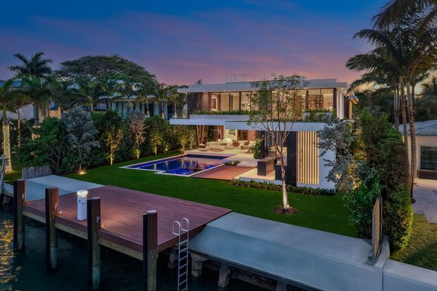 A modern multi-level waterfront residence at dusk, viewed from a timber jetty. The home features extensive glazing, a flat roof, a large covered outdoor living area, landscaped garden, illuminated rectangular swimming pool, and a broad timber deck beside the water. Tall palms and dense tropical planting surround the property.