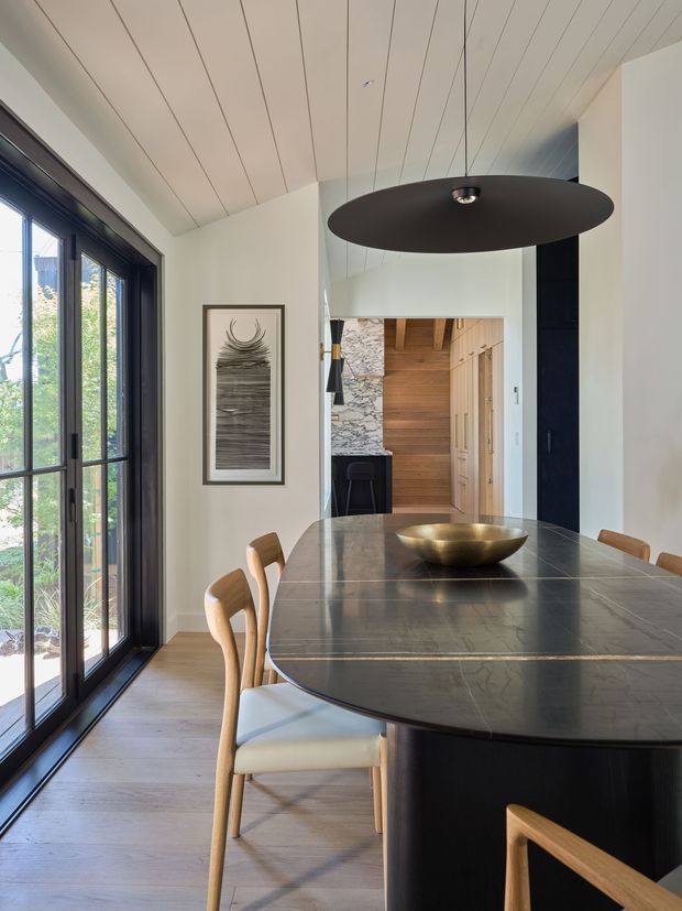 A refined dining area with a large dark stone dining table and rounded corners, surrounded by light timber dining chairs. Full-height dark-framed glazed doors line the left side, opening the room to greenery outside. The space has a white painted-plank ceiling, pale walls, warm timber flooring, a large black disc pendant light, and a sculptural artwork. A hallway or kitchen area with timber joinery and a stone feature wall is visible beyond.