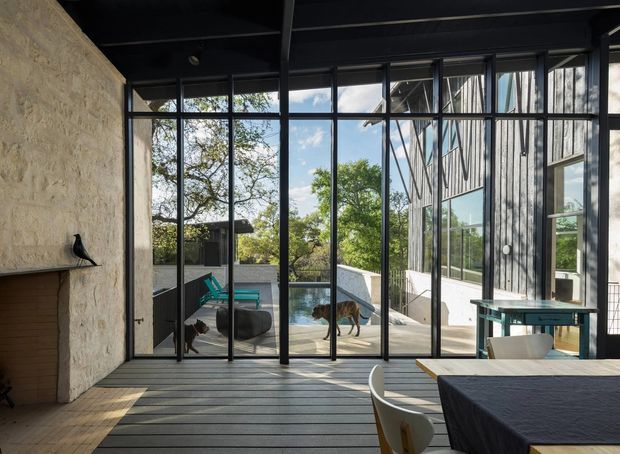 A refined indoor-outdoor living and dining space with dramatic floor-to-ceiling black-framed glazing opening onto a compact pool courtyard. The interior features a dark timber floor, textured pale stone fireplace wall, exposed dark ceiling structure, a dining table with white chairs, and a turquoise console or bar table. Strong natural light and garden views create a contemporary connection to the outdoors.
