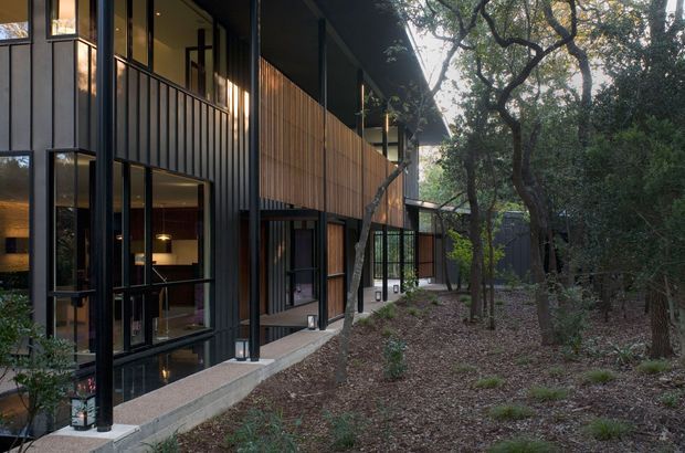 A contemporary multi-level residential building set within a wooded landscape. The facade combines dark vertical cladding, extensive floor-to-ceiling glazing, projecting balconies with slim dark railings, and warm timber screening. The building steps along the site beneath a broad overhanging roof, with mature trees and a natural landscaped foreground.