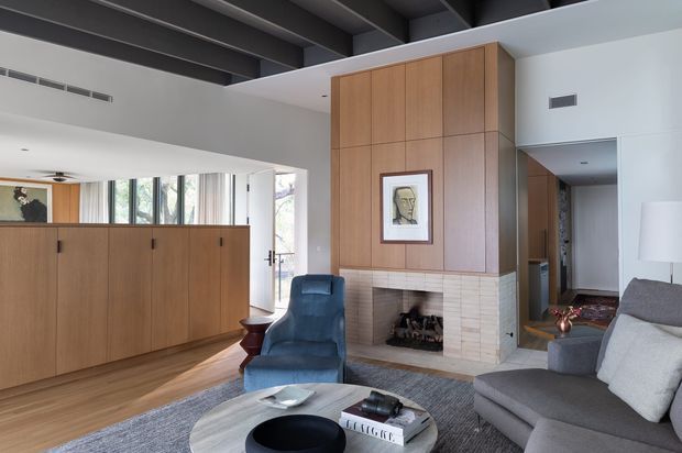 A sophisticated open-plan living room with a large grey sectional sofa, blue lounge chair, round coffee table, and a central fireplace integrated into a tall timber-panelled feature wall. The room has warm timber flooring and cabinetry, a pale tiled fireplace surround, exposed dark ceiling beams, and generous glazing bringing in natural light. A dining or kitchen area is partially visible beyond the timber divider.