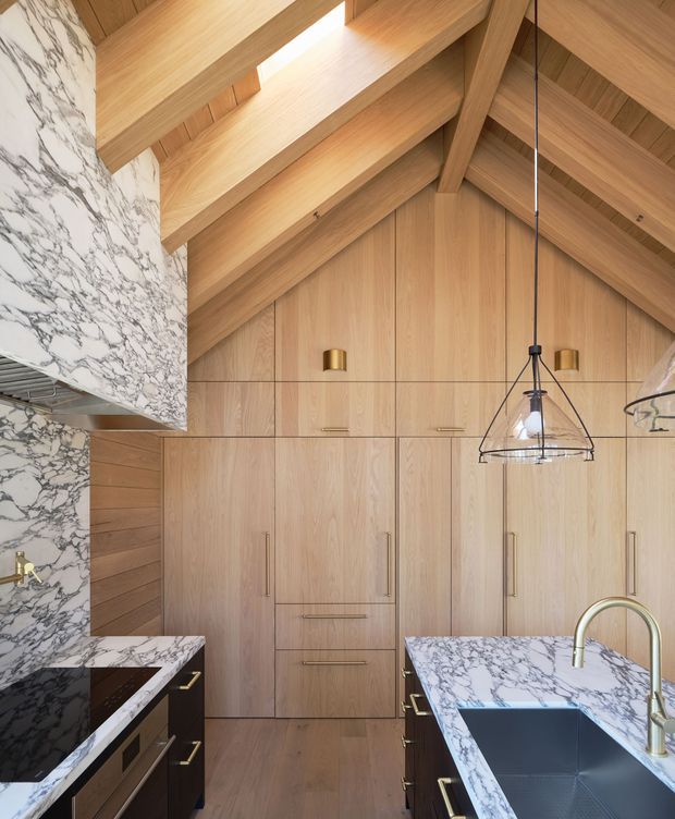 A sophisticated kitchen beneath a dramatic pitched timber-lined ceiling. Full-height light timber cabinetry forms the back wall, complemented by dark lower cabinetry with brass hardware. White marble with prominent grey veining is used on the island and a large overhead or side feature surface. The island includes a deep dark sink with a curved brass tap, while a black geometric pendant light hangs centrally. Warm natural light highlights the timber joinery and exposed roof structure.