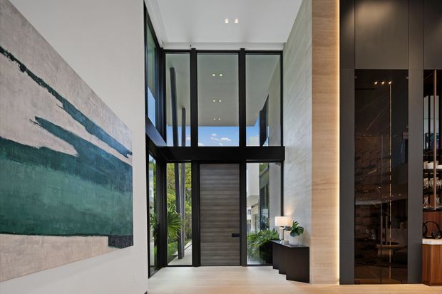 A refined contemporary entry hall with a dramatic double-height glazed void. Black-framed floor-to-ceiling glazing and a dark timber entry door sit between pale textured walls, while a large abstract teal artwork, sculptural planter, console, and integrated dark cabinetry add contrast. A glimpse of the adjoining living area is visible at right.