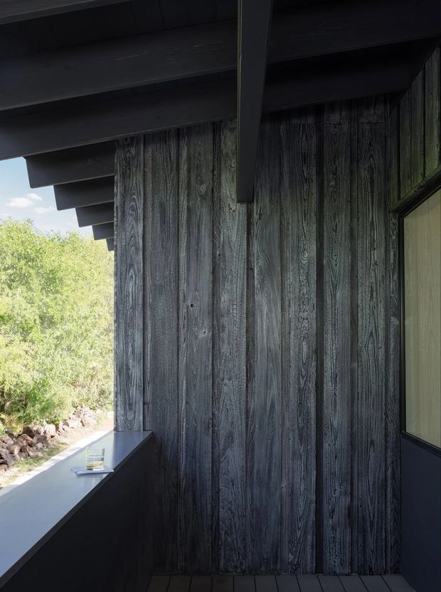 A close architectural exterior detail showing a dark charred or weathered timber-clad wall beneath deep dark timber roof eaves. A narrow projecting ledge or bench runs along the wall beside a glazed opening, with a rocky planted landscape visible beyond. The composition emphasizes strong vertical timber grain, heavy overhangs, and a dark, textural material palette.