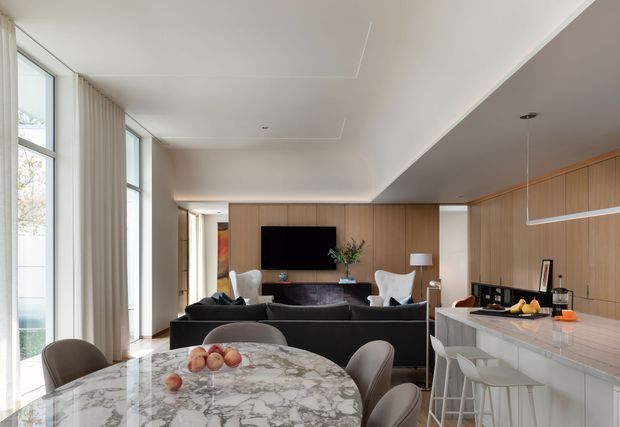 A refined open-plan interior combining a dining area, living room, and kitchen. The foreground features a large round marble dining table with upholstered chairs, while the rear living area has a dark sofa, sculptural white lounge chairs, and a wall-mounted television set against warm timber panelling. The kitchen includes extensive timber cabinetry, a pale stone island with bar seating, and a linear pendant light. Full-height glazing and sheer curtains bring in abundant natural light.
