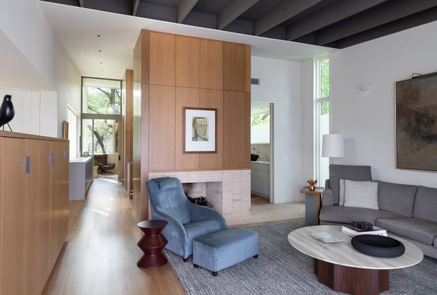 Contemporary open-plan living room with a grey sectional sofa, blue upholstered armchair and ottoman, round coffee table, and textured area rug. A full-height timber-panelled fireplace wall with pale brick surround forms the focal point, while built-in timber cabinetry lines the adjacent hallway. Exposed dark ceiling beams contrast with white walls, and large windows bring in abundant natural light and views of greenery.