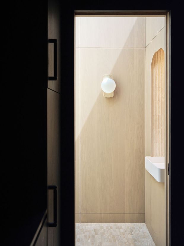 A narrow, dimly lit bathroom or powder-room interior with pale timber wall panelling, a concealed door or cabinetry front, a small spherical wall light, and a partially visible white basin set into a side niche. The floor is finished in small pale stone or tile pieces, and an arched timber-lined opening adds a subtle feature detail. The image has a minimalist, warm, highly controlled composition with strong contrast between the dark surrounding walls and the light timber interior.