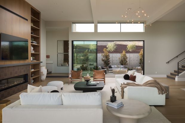 A spacious contemporary living room with high ceilings, large clerestory windows and expansive glazing opening toward a landscaped courtyard. The room features white sofas, a rounded coffee table, a dark oval coffee table, a built-in timber entertainment wall with open shelving, a brick fireplace, and a modern sculptural pendant light. A staircase is visible to the right, while the courtyard is seen through the glazing.
