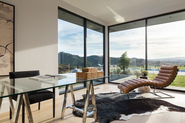 Bright contemporary open-plan dining and lounge area with floor-to-ceiling corner glazing overlooking a landscaped setting and distant hills. A glass dining table with black chairs occupies the foreground, while a brown leather chaise lounge and dark hide rug create a relaxed seating area. The space has a light neutral palette, clean lines, and abundant natural light.
