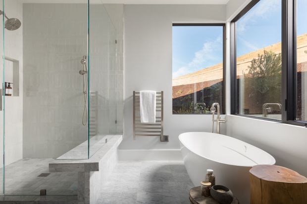 Bright contemporary bathroom with a large frameless glass walk-in shower, freestanding white oval bathtub, wall-mounted chrome fixtures, pale stone or concrete-look surfaces, and a timber towel ladder. Large black-framed windows bring in natural light and overlook an exterior courtyard wall. A timber stool and small decorative accessories add warmth.