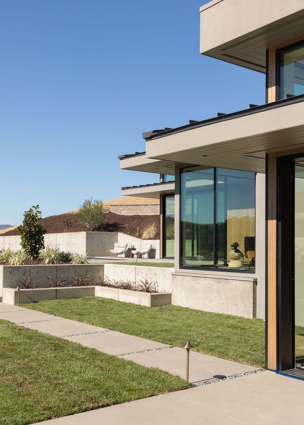 Exterior view of a contemporary multi-level residence with strong horizontal projecting roof slabs, expansive floor-to-ceiling glazing, dark window frames, and pale rendered or concrete walls. The home opens onto a landscaped garden with lawns, concrete paths, raised planting beds, and a small outdoor seating area. Clear blue sky and layered roof forms emphasize the crisp modern design.