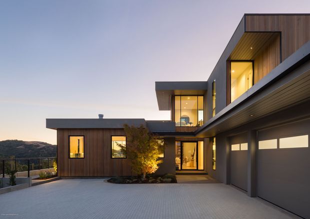 Contemporary multi-level residential home photographed at dusk, with dark timber cladding, charcoal metal rooflines and fascia, extensive glazed openings, warm interior lighting, and an integrated double garage. A broad paved driveway and landscaped entry courtyard occupy the foreground, while a hilly landscape is visible beyond the property.
