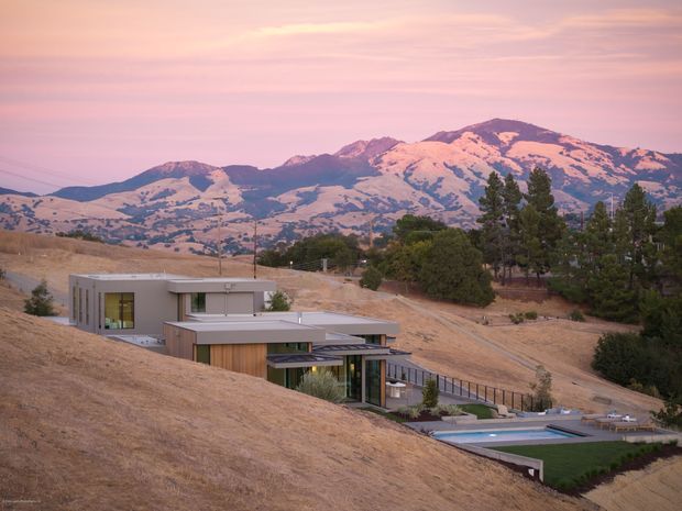 Wide exterior view of a contemporary, low-profile residence embedded into a dry, sloping hillside. The home has crisp rectangular forms, flat roofs, extensive glazing, and warm timber-toned cladding, with a pool terrace visible beside the house. A rugged mountain range dominates the background beneath a pink sunset sky.