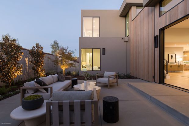 Twilight view of a contemporary multi-storey home with a landscaped courtyard patio and outdoor lounge furniture. The facade combines smooth rendered walls with vertical timber cladding and expansive glazed openings. Warm interior lighting is visible through the open glass door, while the patio includes a sectional sofa, armchairs, low tables, and architectural planting.