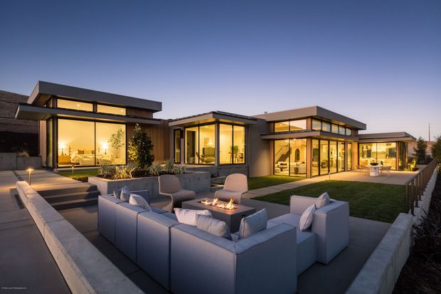 Twilight view of a low-profile modern residence arranged around a landscaped central courtyard. The composition features extensive floor-to-ceiling glazing, flat rooflines, dark and warm neutral exterior finishes, and a large outdoor lounge with sectional seating surrounding a built-in fire feature. Interior lighting creates a warm glow through the windows, while paved terraces and clipped lawn define the outdoor living area.