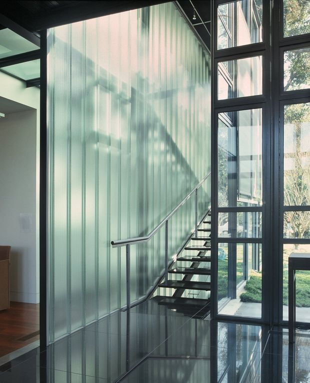 A contemporary interior stairway with a floating open-tread staircase, slender metal handrail, translucent vertically ribbed glass wall, dark metal-framed floor-to-ceiling glazing, and a glossy dark floor reflecting the structure. A landscaped courtyard is visible through the windows, with part of an adjoining living area at left.