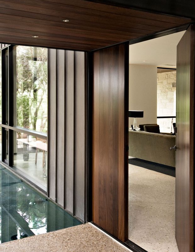 Interior architectural view of a refined modern hallway or entry passage beside a narrow indoor reflecting pool or water feature. The space features dark timber ceiling boards and wall panels, slim black-framed floor-to-ceiling glazing, a terrazzo-like speckled floor, and a pivot door opening toward a light-filled living area with a stone feature wall and sofa. Strong connections to surrounding greenery are visible through the glazing.