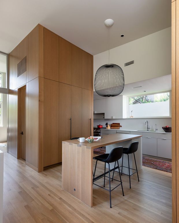 A warm, contemporary kitchen viewed from an adjoining circulation area. Full-height natural timber cabinetry forms a substantial storage wall, while a timber island with an integrated dining overhang is paired with black bar stools. The kitchen has pale cabinetry, a light timber floor, a large horizontal window, stainless-steel tapware, and a prominent woven pendant light. A hallway and glazed opening are visible to the left.