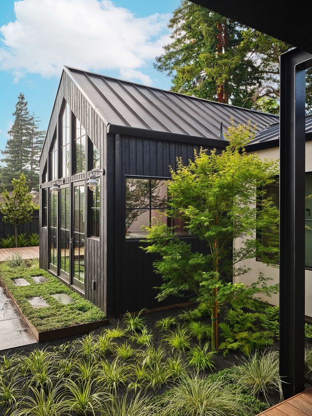 A contemporary dark-clad residential extension or pavilion set within a landscaped garden. The building has steeply pitched gable roofs with standing-seam metal roofing, black vertical timber cladding, expansive black-framed glazing, and a paved garden path bordered by ornamental grasses and leafy planting. A covered outdoor structure frames the right edge of the image, while mature trees and blue sky form the background.