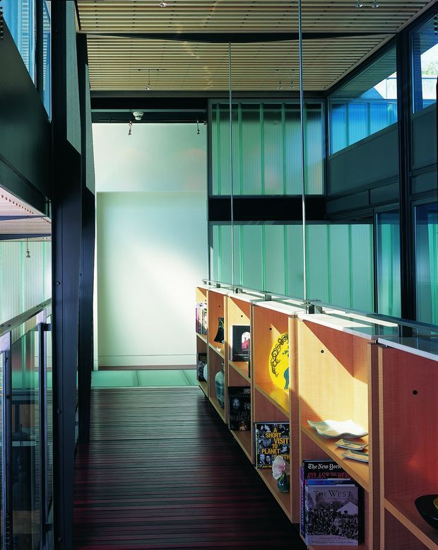 A narrow, multi-level interior walkway or gallery-like corridor with dark timber flooring, black structural framing, extensive blue-green glazed partitions, and a slatted timber ceiling. Built-in timber display shelving lines the right side, holding books and decorative objects, while strong daylight filters through the glazing. The space has a contemporary, industrial-leaning architectural character with a tall, open vertical volume.