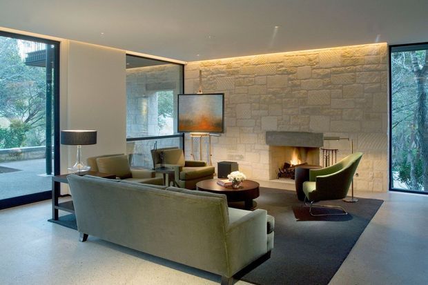 A refined contemporary living room with floor-to-ceiling glazing, a large textured stone feature wall and built-in fireplace. The seating arrangement includes a muted green sofa, olive lounge chairs, a dark round coffee table, floor lamp and rug, with warm concealed lighting washing across the stonework. Surrounding trees are visible through the windows.