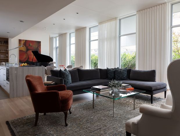 Bright, open-plan contemporary living room with a long charcoal sectional sofa, sculptural lounge chairs, glass coffee table, textured area rug, and a black grand piano. Floor-to-ceiling windows with vertical sheer blinds bring in abundant natural light. The adjacent kitchen features pale wood-grain cabinetry and a stone or light benchtop, while a colourful abstract artwork adds visual contrast.