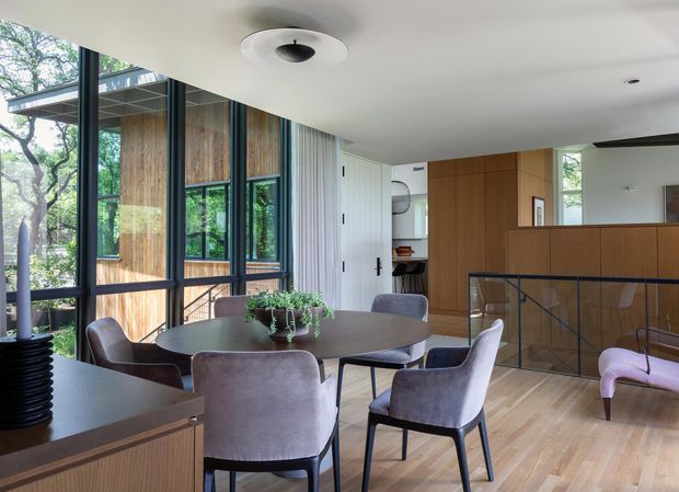A refined open-plan dining and living area with a round dark dining table surrounded by upholstered chairs, set beside extensive black-framed glazing overlooking leafy trees. Warm timber wall panelling and cabinetry, pale timber flooring, and a minimalist white ceiling create a calm contemporary interior. A glazed stair or balustrade area and adjoining kitchen/living zone are visible in the background.