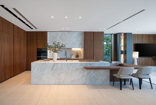 A refined open-plan kitchen and living area with a large veined stone island and matching splashback, extensive warm timber cabinetry, integrated appliances, and a built-in dining extension. Pale timber flooring, recessed linear ceiling lighting, upholstered bar stools, and a television lounge visible to the right create a clean, contemporary interior.