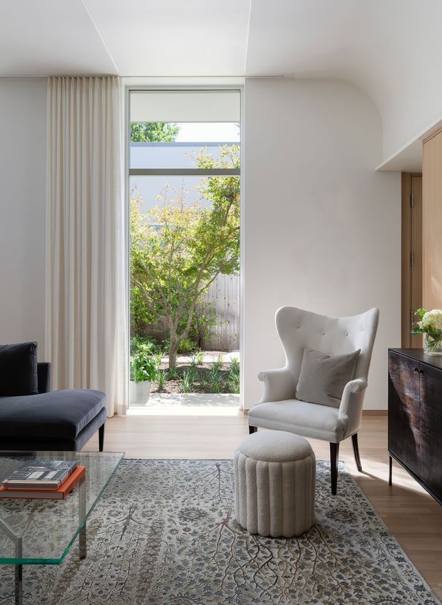Bright, refined living room with a tall glazed garden door or window, sheer floor-to-ceiling curtains, pale timber flooring, a patterned area rug, a dark sectional sofa, and a tufted wingback armchair with matching ottoman. A glass coffee table and dark timber sideboard complete the calm contemporary interior. The room opens visually to a small planted courtyard or garden.
