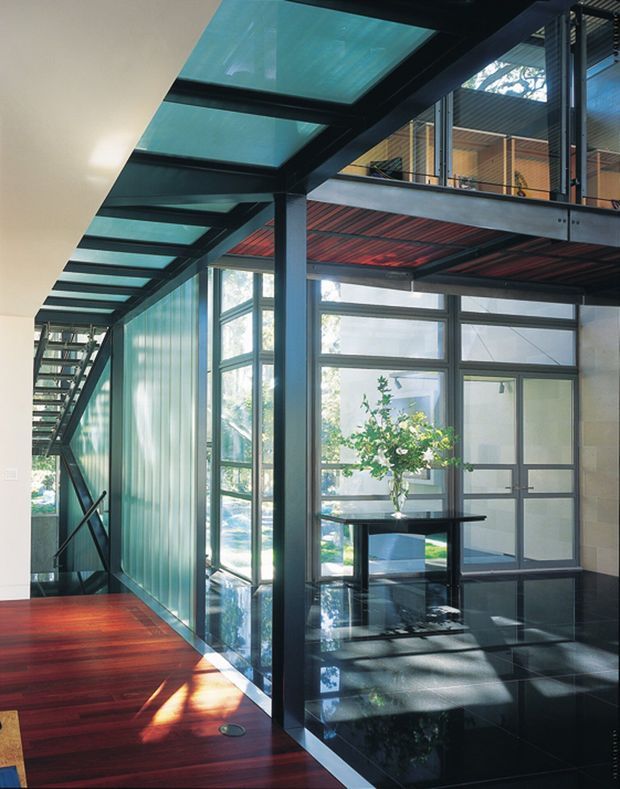A dramatic modern interior atrium or circulation space with extensive floor-to-ceiling glazing, a black steel-framed glass roof, exposed structural steel, and an upper-level mezzanine accessed by an open stair. The space combines glossy black tiled flooring with warm reddish timber flooring, translucent ribbed wall panels, and abundant natural light. A minimalist console table with a vase sits near the glazed entrance area.