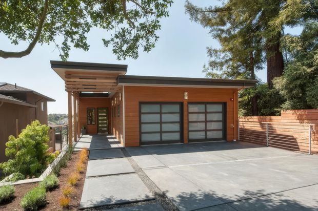Contemporary single-storey residential garage and entry facade with warm horizontal timber cladding, two large frosted-glass garage doors, a covered timber-framed entry walkway, and a broad concrete driveway. Mature trees and a horizontal timber boundary fence frame the property.
