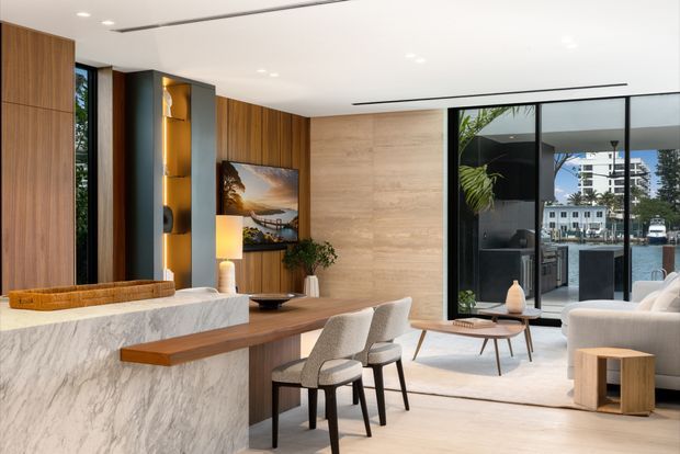 A refined open-plan living and dining space with a marble kitchen island and integrated timber breakfast bar in the foreground. Vertical timber wall panelling, warm recessed display shelving, a large artwork, contemporary dining chairs, a low coffee table, and a pale upholstered sofa create a warm modern interior. Full-height black-framed glazing opens views toward an outdoor terrace and water beyond.