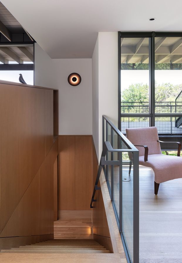 Interior view of a contemporary upper-level landing and stairway. A broad timber-lined built-in wall and stair surround runs along the left, while a glass balustrade with dark metal framing borders the stair opening. Tall black-framed windows bring in abundant daylight and overlook leafy surroundings. A sculptural wall sconce and upholstered armchair add refined accents to the otherwise restrained space.