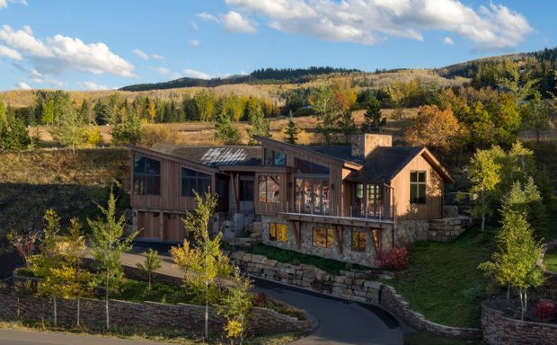 A rustic contemporary mountain home set into a landscaped, sloping site. The single-storey timber-clad dwelling has several interconnected pitched roof volumes, broad glazed openings, a raised timber deck, stone retaining walls, and a curved driveway. Autumnal woodland and rolling hills surround the property.