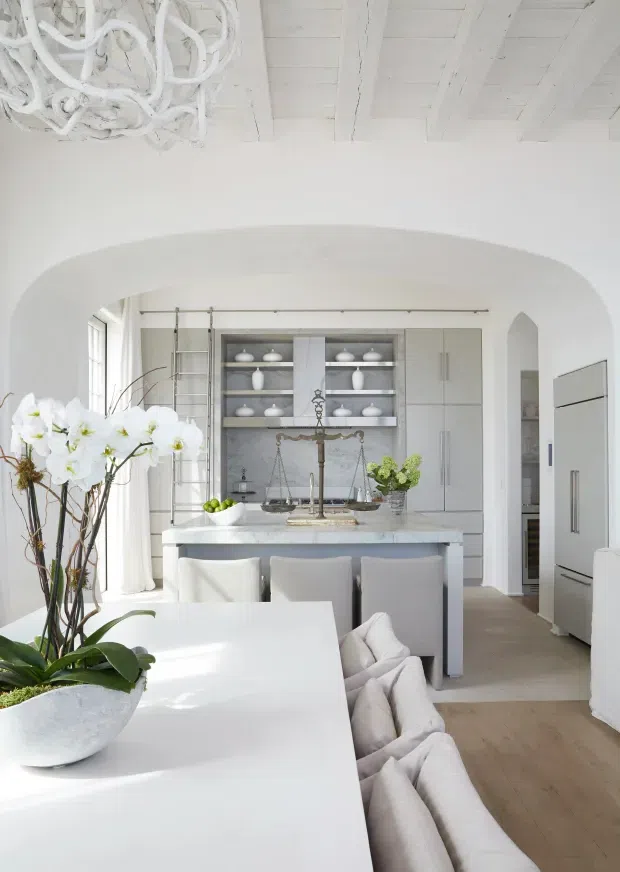 A bright, refined open-plan kitchen and dining area in a predominantly white interior. The scene features a large white dining table with upholstered seating, a central island with a pale stone benchtop, white cabinetry, open shelving displaying ceramics, integrated appliances, and a decorative antique-style brass tap and scale. White-painted ceiling beams, a broad arched opening, a sculptural white chandelier, light timber flooring, and an orchid arrangement complete the airy design.