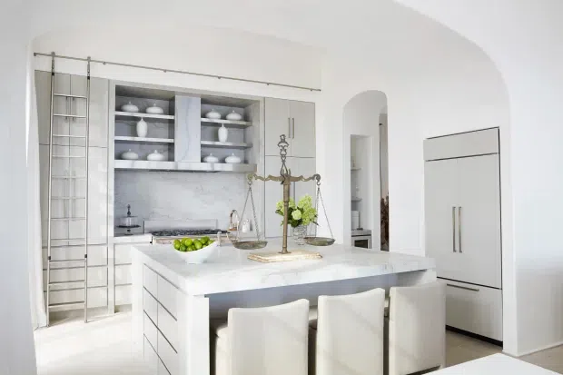 Bright, refined white kitchen with a large marble-look island, integrated cabinetry, open display shelving, a tall rolling library ladder, and three upholstered island stools. Arched openings and concealed appliances contribute to a polished, classic interior aesthetic.