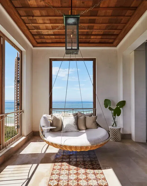 Bright coastal sitting area with a suspended circular rattan lounge chair positioned beside large ocean-facing windows. The room features white plaster walls, warm timber ceiling panelling with exposed beams, dark timber window frames, tiled flooring, patterned runner rug, and a potted plant. Strong sunlight creates a warm, airy atmosphere with expansive sea views.