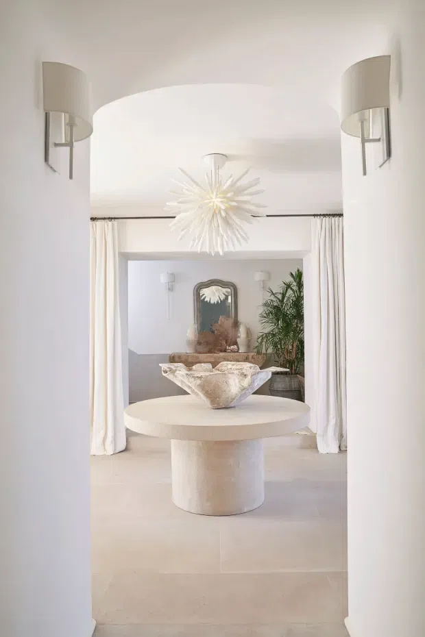 A refined, light-filled interior view through a curved white archway into a formal entry or living area. A round cream stone pedestal table with a sculptural vessel occupies the foreground, beneath a dramatic white starburst pendant. Full-height white curtains frame the rear opening, while curved wall sconces, a decorative mirror, neutral furnishings, and indoor greenery create a soft, luxurious atmosphere.