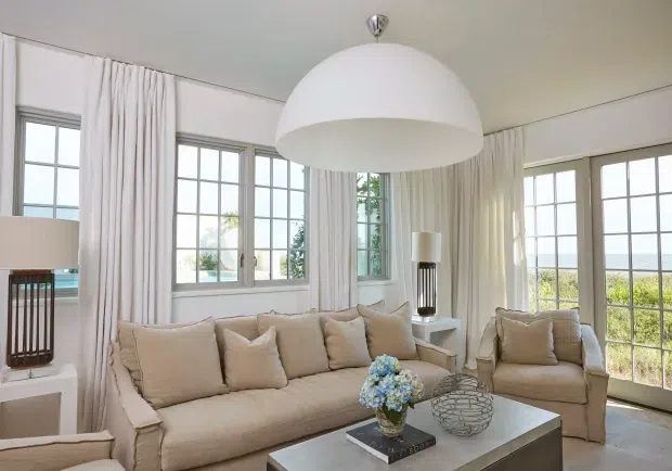 Bright, elegant living room with a large beige upholstered sofa and matching armchair arranged around a light rectangular coffee table. Tall multi-pane windows and full-height sheer curtains provide abundant natural light, while a prominent white dome pendant light hangs overhead. The room has a refined neutral palette with subtle decorative accessories and views of greenery and water beyond the glazing.