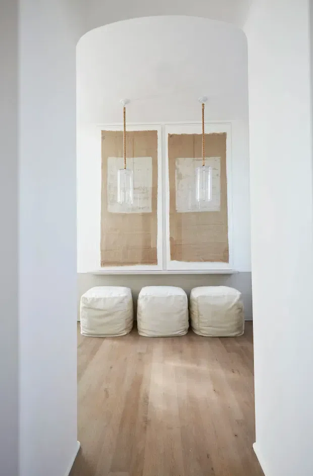 A bright, minimalist interior alcove viewed through a wide rounded archway. Three white upholstered poufs sit along the back wall beneath two framed neutral-toned artworks and delicate hanging pendant lights. Pale timber flooring, white walls, and a soft grey lower wall create a calm, understated composition.