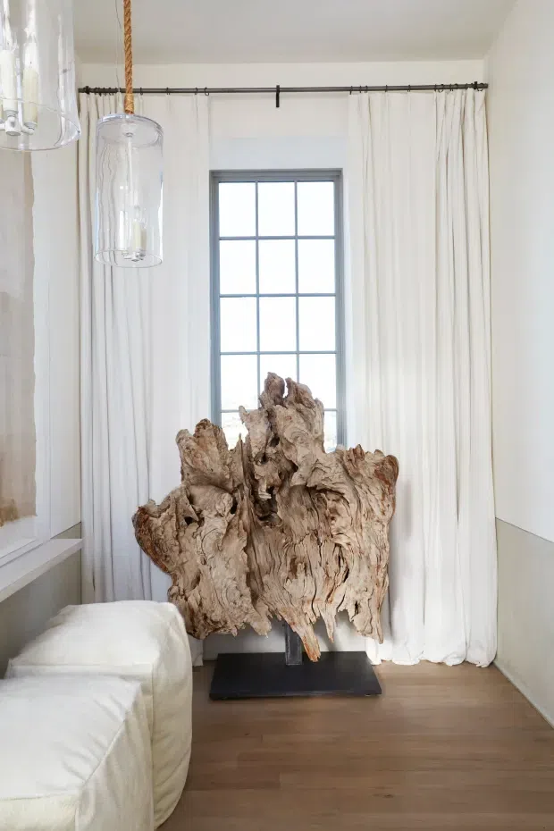 Bright, serene interior corner with a large sculptural natural timber root displayed on a black plinth in front of a tall multi-pane window. Sheer white curtains span the wall, while a white upholstered sofa or bed edge appears at left. Clear glass pendant lights with rope cords hang overhead, and the room has pale walls and warm timber flooring.