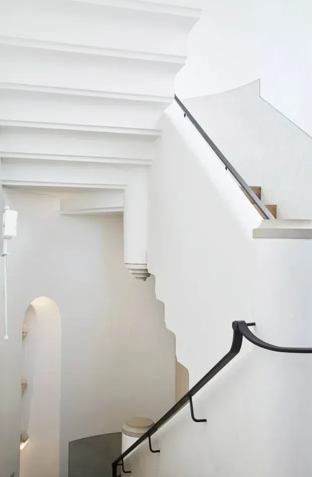 Bright, predominantly white interior stairwell with a sculptural staircase, black metal handrails, and pale stone or concrete stair treads. Layered ceiling and wall mouldings frame the stair opening, while a softly arched doorway and minimalist furnishings are visible below. The image emphasizes the vertical circulation space and refined monochromatic detailing.