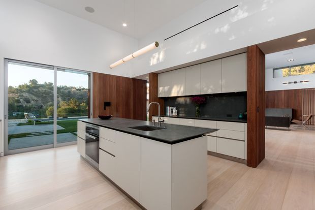 A bright, open-plan contemporary kitchen with a large central island, white handleless cabinetry, dark stone benchtops, and a glossy dark splashback. Warm timber wall panels and structural-looking columns frame the kitchen and connect it to an adjacent living area. Pale timber flooring, white walls, linear pendant lighting, and full-height sliding glazing create an airy indoor-outdoor connection, with landscaped grounds visible beyond.