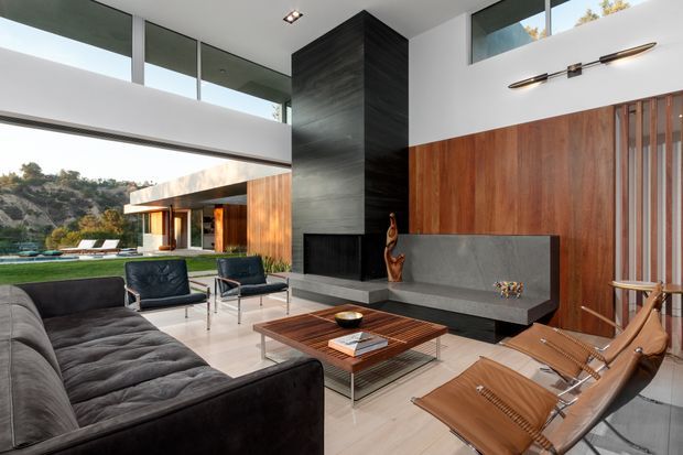 A spacious modern living room with floor-to-ceiling glazing, high clerestory windows, a dramatic dark fireplace volume, and warm timber wall panelling. The room includes a large charcoal sectional sofa, leather lounge chairs, a low timber coffee table, and a built-in grey fireplace bench. The glazing opens views toward a landscaped outdoor area and rocky terrain, while a linear wall light and recessed ceiling lighting provide understated illumination.