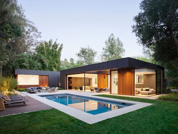 A single-storey modern courtyard-style residence with a flat roof, dark exterior cladding and extensive floor-to-ceiling glazing arranged around a rectangular swimming pool. Warm timber-lined walls and soffits create contrast, while an adjacent deck includes outdoor lounge furniture and sun loungers. The landscaped garden and mature trees surround the contemporary pool pavilion.